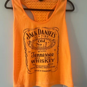 Orange topp - Säljer en orange linnetopp med stort svart Jack Daniel's Tennessee Whiskey-tryck framtill. Toppen är croppad och har brottarrygg i meshmaterial som ger en sportig känsla. Perfekt för varma dagar eller festival. Oanvänd