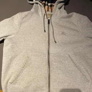 Grå zip-hoodie från Burberry (äkta) - Säljer en grå hoodie från Burberry med dragkedja och klassiskt rutigt foder i huvan och längst insidan. Den har fickor framtill och justerbar huva med snörning. Perfekt för dig som gillar stilrena plagg med en twist. Går att diskutera pris vid snabb affär 