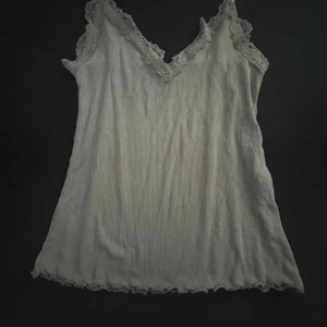 Vit ribbad linnetopp med spets från Amisu - Säljer en söt vit ribbad linnetopp från Amisu med spetsdetaljer längs v-ringningen och axelbanden. Perfekt att styla till jeans eller kjol för en fräsch look.