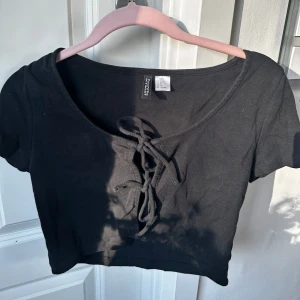 Svart croppad topp med snörning från H&M Divided - Säljer en svart croppad topp från H&M Divided med korta ärmar och snygg snörningsdetalj framtill. Perfekt till jeans eller kjol för en trendig look. Topp i mjukt bomullsmaterial. ❤️