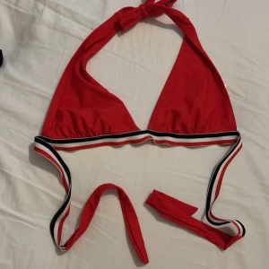 Röd bikiniöverdel med knyt - Snygg röd bikiniöverdel med knyt i nacken och under bysten. Har sportiga ränder i svart, vitt och rött längs kanten. Perfekt för stranden eller poolen!