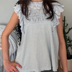 Blå blus med spets och volang - Supersöt blå blus med spetsdetaljer och volanger på axlarna. Blusen har ärmlös design och vackert broderat mönster upptill.  Storlek M men passar S.
