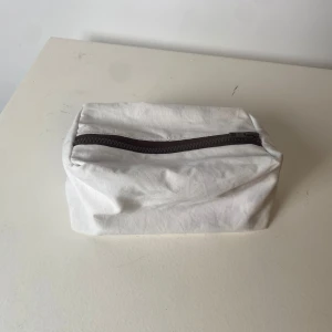 Vit necessär med dragkedja - En enkel och stilren vit necessär med svart dragkedja upptill. Perfekt att ha med sig för att organisera småsaker eller smink. Rektangulär form och tillverkad i ett mjukt material.