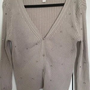 Beige ribbad kofta med pärlor från H&M - Säljer en beige ribbad kofta från H&M med v-ringning och knäppning framtill. Koftan har långa ärmar och är dekorerad med små pärlor över hela plagget för en extra detalj. Perfekt att slänga över en topp eller klänning.