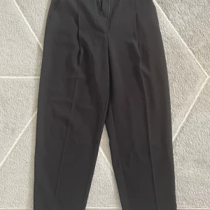 Svarta kostymbyxor med pressveck - Säljer ett par klassiska svarta kostymbyxor med pressveck och hällor för bälte. Byxorna har normal passform och stängs med dragkedja och hake. Perfekta till en stilren look och passar till många olika tillfällen.