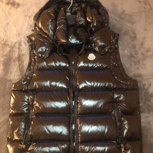 Svart dunväst från Moncler | ev byte - Säljer en svart, glansig dunväst från Moncler med avtagbar huva och dragkedja framtill. Västen har två fickor med dragkedja och Moncler-logga på bröstet. Perfekt för lager-på-lager under kyliga dagar.