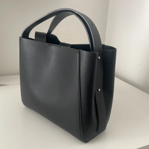 Svart handväska i skinn - Stilren svart handväska med minimalistisk design och breda handtag. Väskan är tillverkad i slätt skinn och har silverfärgade nitar på sidorna. Perfekt för dig som gillar en clean och modern look.