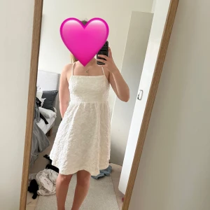 Vit spetsklänning med öppen rygg och smala axelband - Superfin vit klänning med smala axelband och öppen rygg, justerbar med snören. Klänningen har en lätt utställd kjol och är täckt av vacker spets med blommigt mönster. 