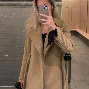 Beige kappa med klassisk krage - Stilren beige kappa med klassisk krage och snygg passform. Perfekt för dig som vill ha en tidlös jacka till garderoben. Jackan har långa ärmar och stängs med knappar framtill.