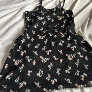 Svart blommig klänning med smala axelband - Säljer en svart klänning med små blommor i rosa och vitt. Klänningen har smala axelband och en lätt utställd passform. Perfekt för varma dagar eller att styla med en tröja under.