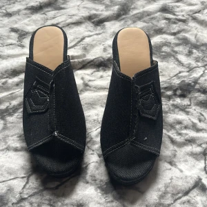 Svarta kilklackar i jeansmaterial - Säljer ett par svarta sandaler med kilklack i flätad stil och ovandel i jeansmaterial. Skorna har öppen tå och en dekorativ fickdetalj framtill. Perfekta för dig som gillar en unik och trendig look.