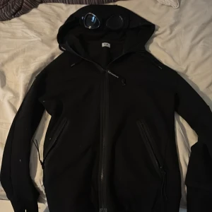 Svart hoodie från C.P. Company - Säljer en svart hoodie från C.P. Company med ikoniska goggles i huvan. Hoodie har dragkedja framtill och två snedställda fickor med dragkedja. Perfekt för dig som gillar streetwear och vill sticka ut.