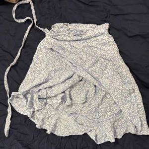 Ljusblå blommig omlottkjol med volang - Supersöt ljusblå omlottkjol med små vita blommor och volangdetaljer längs kanten. Kjolen knyts i midjan och har en luftig känsla, perfekt för varmare dagar.