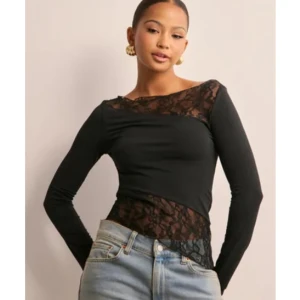 Svart topp med spetsdetaljer - Snygg svart långärmad topp med asymmetrisk spetsdetalj upptill och nedtill. Toppen är endast använd 1 gång! Den är i storlek S. Toppen är från Nelly och finns inte kvar i deras sortiment 