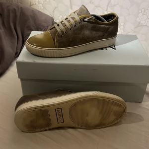 Grå sneakers från Lanvin med beige detaljer - Säljer ett par stilrena grå sneakers från Lanvin med beige tåparti och vita sulor. Skorna har snörning och är tillverkade i mocka med detaljer i skinn. Storlek är 39 men passar även 40, 41. Skicket är 8/10. Box ingår ej endast dustbag om ni har några funderingar tveka inte att höra av er angående skorna.