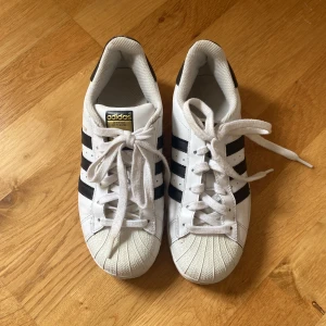 Adidas Superstar vita sneakers med svarta ränder - Klassiska Adidas Superstar sneakers i vitt med svarta ränder och snörning. Skorna har rund tå och platt sula, samt den ikoniska snäckformade tån. Perfekta för en sportig och stilren look.