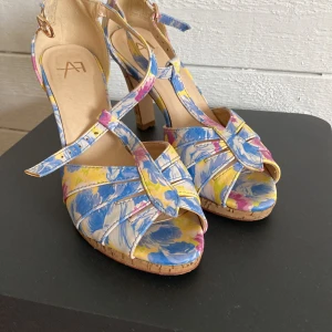 Blommiga klackskor med korkklack från AF - Färgglada klackskor med blommigt mönster i blått, gult, rosa och vitt. Skorna har öppen tå, knytning framtill och hög korkklack. Perfekta för dig som vill sticka ut med en somrig och lekfull stil.