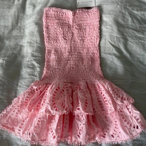 Rosa klänning från Design by Amor - Supersöt rosa klänning från Design by Amor med smock upptill och volanger nertill. Klänningen är helt oanvänd och lappen sitter även kvar. Köparen står för frakten 😊
