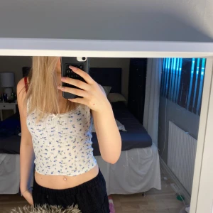 Vit blommig linnetopp med spetsdetaljer - Säljer en söt vit linnetopp med små blå blommor och gröna blad. Toppen har smala axelband och spets längs kanterna. Perfekt för varma dagar och enkel att matcha med jeans eller kjol.