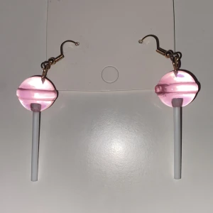 Rosa klubba-örhängen INGA PLUPPAR - Säljer ett par unika örhängen formade som rosa klubbor med vita pinnar. Perfekta för dig som vill sticka ut och addera en lekfull touch till din stil. Hängande modell med guldfärgade krokar.