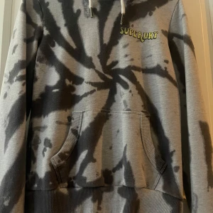Superdry hoodie - Säljer en grå och svart tie-dye hoodie från Superdry med tryckt logga fram och svampar i gult på ryggen. Tröjan har huva med dragsko och magficka. Perfekt för dig som gillar unika detaljer!