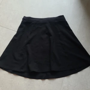 Svart kortkjol från H&M - Säljer en svart kortkjol från Divided med en dragkedja baktill. Kjolen har en lätt utställd form och är tillverkad i ett mjukt bomullsmaterial. Perfekt att styla med en enkel topp.