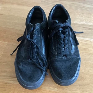Svarta Vans sneakers - Klassiska svarta Vans sneakers med snörning och rund tå. Skorna är i mocka och syntet, med platt sula och diskret Vans-logga på sidan. Perfekta för en avslappnad och stilren look.