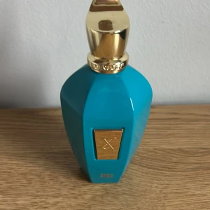 Turkos parfymflaska från Xerjoff - Exklusiv parfym från Xerjoff i en turkos flaska med gulddetaljer och elegant lock. Flaskan har en geometrisk form och en metallplatta framtill med märkets logotyp. Perfekt för dig som gillar lyxiga dofter och snygg design.