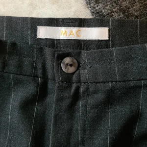 Sjukt stiliga kostymbyxor från MAC! - Snygga svarta kostymbyxor från MAC med tunna vita ränder. Klassisk passform med raka ben och knappstängning i midjan. Långa och stiliga med pressveck.