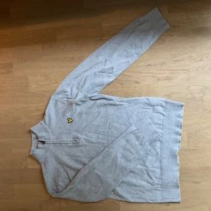 Grå halvzip tröja från Lyle & Scott - Säljer en ljusgrå halvzip tröja från Lyle & Scott med deras klassiska gula fågel-logga broderad på bröstet. Tröjan har lång ärm och hög krage med dragkedja. Perfekt för lager-på-lager och enkel att matcha.