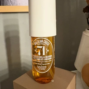 Brazilian Crush Cheirosa 71 Hair & Body Fragrance Mist - En söt och varm doftmist från Sol de Janeiro med toner av karamelliserad vanilj och macadamia. Flaskan är genomskinlig med gyllene vätska och har en stor vit kork. Perfekt för både hår och kropp.