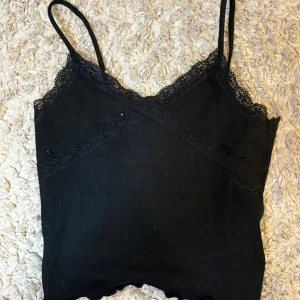 Svart ribbad linnetopp med spets - Säljer en svart ribbad linnetopp med smala axelband och fina spetsdetaljer längs urringningen. Perfekt att styla under en skjorta eller på egen hand för en enkel och snygg look.