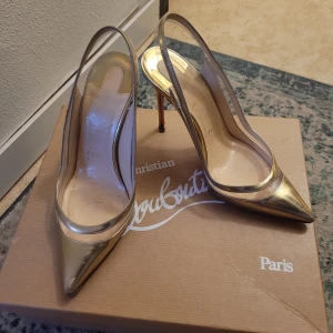 Guldiga klackar från Christian Louboutin - Säljer ett par eleganta guldiga klackar från Christian Louboutin med spetsig tå och hög klack på 100mm. Skorna har transparenta detaljer på sidorna och klassisk röd sula. Perfekta för dig som vill sticka ut med lyxig stil. Box och nya "klacktippar" följer med🩷 självklart äkta!