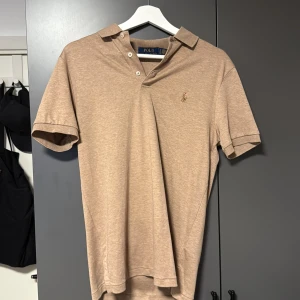 Beige pikétröja från Polo Ralph Lauren - Säljer en klassisk beige pikétröja från Polo Ralph Lauren med korta ärmar och krage. Tröjan har två knappar vid halsen och den ikoniska broderade loggan på bröstet. Perfekt för en stilren och avslappnad look. Aldrig använd STORLEK S
