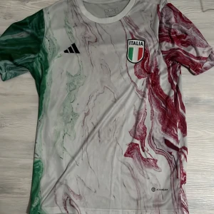 Italien fotbollströja Adidas - Säljer en snygg Italien fotbollströja från Adidas med marmorerat mönster i grönt, vitt och rött. Tröjan har korta ärmar, broderad Italien-logga och Adidas-märke på bröstet. Perfekt för dig som vill sticka ut på match eller träning.