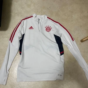 Vit långärmad Bayern München träningströja från Adidas - Säljer en vit långärmad träningströja från Adidas med Bayern München-logga och röda detaljer. Tröjan har dragkedja vid halsen, röda ränder på ärmarna och mörkblå partier på sidorna. Perfekt för fotbollsträning eller support.