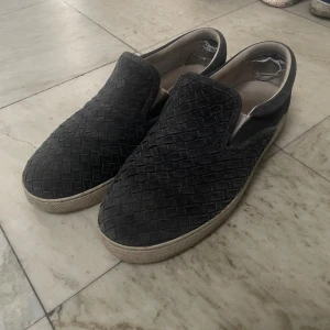 Svarta slip-on sneakers med flätad ovandel - Säljer ett par svarta slip-on sneakers med snygg flätad ovandel och vit sula. Skorna har rund tå och är perfekta för en avslappnad stil. Passar både till jeans och shorts.