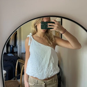 Vit broderad linnetopp från Zara - Säljer en söt vit linnetopp från Zara med broderade detaljer och knyt på ena axeln. Toppen är croppad och har en luftig känsla, perfekt för varma dagar.