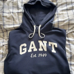 Mörkblå hoodie från GANT - Säljer en mörkblå hoodie från GANT med vit logga och texten 'Est. 1949' på bröstet. Tröjan har huva med dragsko och är perfekt för en avslappnad stil.