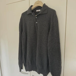 Mörkgrå stickad pikétröja från Gran Sasso - Mörkgrå stickad pikétröja från Gran Sasso i en lyxig blandning av ull och kashmir. Tröjan har klassisk krage, knäppning med tre knappar och långa ärmar. Perfekt för dig som gillar stilrena och bekväma plagg. Priset är inte hugget i sten🪨