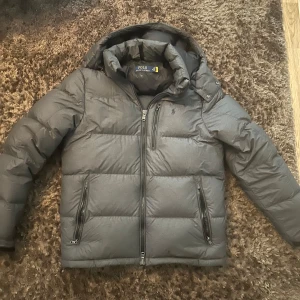 Grå dunjacka från Polo Ralph Lauren - Säljer en grå dunjacka från Polo Ralph Lauren med huva och dragkedjor framtill samt på fickorna. Jackan har en diskret logga på bröstet och är perfekt för kyliga dagar. Klassisk pufferstil med lång ärm och snygg passform.