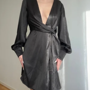 Svart omlottklänning med ballongärmar Nelly  - Snygg svart omlottklänning med djup v-ringning och långa ballongärmar med knappar vid ärmsluten. Klänningen har en markerad midja med knytband och en liten slits framtill. Materialet har en lätt glansig finish som ger en lyxig känsla.