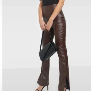 Bruna PU-byxor med slits Nelly Trend  - Snygga bruna PU-byxor med hög midja och slits nertill på benen. Byxorna har en slim passform och är tillverkade i ett glansigt syntetmaterial som ger en cool läderlook. Perfekta att styla med klackar eller boots. Passar s-mindre m