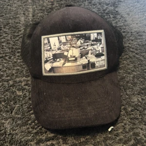 Svart keps från Sayless med tryck - Svart keps från Sayless med justerbar baksida och meshpaneler. Framtill finns ett svartvitt tryck med motiv av ett kontor. Kepsen har broderad logotyp på insidan och svensk flagga på etiketten.