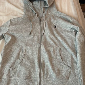 Polo Ralph Lauren zip up - 100% cotton nästan aldrig använd. Snygg grå hoodie från Ralph Lauren med dragkedja och två fickor framtill. Klassisk svart logga broderad på bröstet och justerbar huva med snören. Perfekt för en avslappnad stil.
