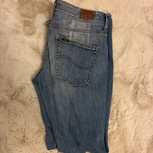 Ljusblå bootcut jeans från Lee - Säljer ett par ljusblå jeans från Lee med klassisk femficksdesign och bootcut passform. Jeansen har knappar i gylfen och subtila slitningar framtill för en avslappnad look. Perfekta för dig som gillar en tidlös stil.