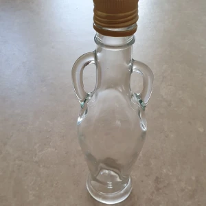 Glasflaska - Säljer en stilren och dekorativ glasflaska med två handtag och skruvkork i beige. Flaskan har en klassisk, amforainspirerad form och passar perfekt som inredningsdetalj eller för servering av dryck.