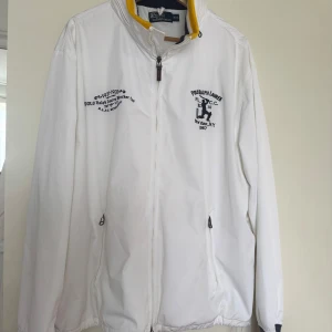 Vit vindjacka från Polo Ralph Lauren - Säljer en vit vindjacka från Polo Ralph Lauren med broderade detaljer på bröstet och dragkedja framtill. Jackan har ståkrage med gul insida och två fickor med dragkedja. Perfekt för dig som gillar sportig och klassisk stil. Även en infälld luva