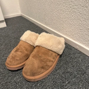 Bruna tofflor med fuskpäls - Mysiga bruna tofflor med mjuk beige fuskpäls runt öppningen. Ovandelen är i mockaliknande material och sulan är platt och mönstrad för bra grepp. Perfekta för att hålla fötterna varma hemma. Storlek 40-41 men skulle säga att de passar 39-40😇