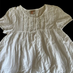 Vit blus med spetsdetaljer från Dreams - Söt vit blus från Dreams i storlek 140. Blusen har korta ärmar, fina spetsdetaljer längs bröstet och nederkanten samt en luftig och lös passform. Perfekt för en somrig och avslappnad look.